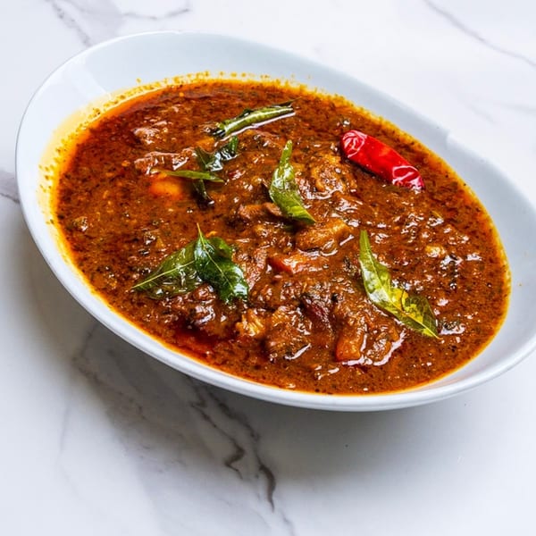 Spicy Chettinad curry with meat, garnished with green leaves and a red chili in a white bowl.