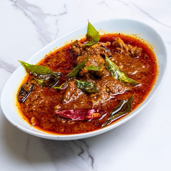 A white oval dish filled with a rich, red Indian curry, garnished with green leaves and red chilies.