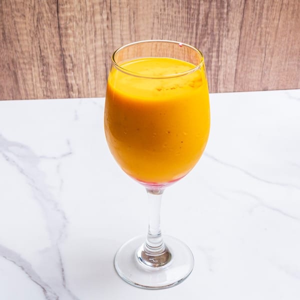 A tall glass of vibrant orange mango lassi sits on a white marble surface.