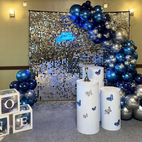 A birthday party setup with blue and silver balloons, a sequin backdrop, and white pedestals.