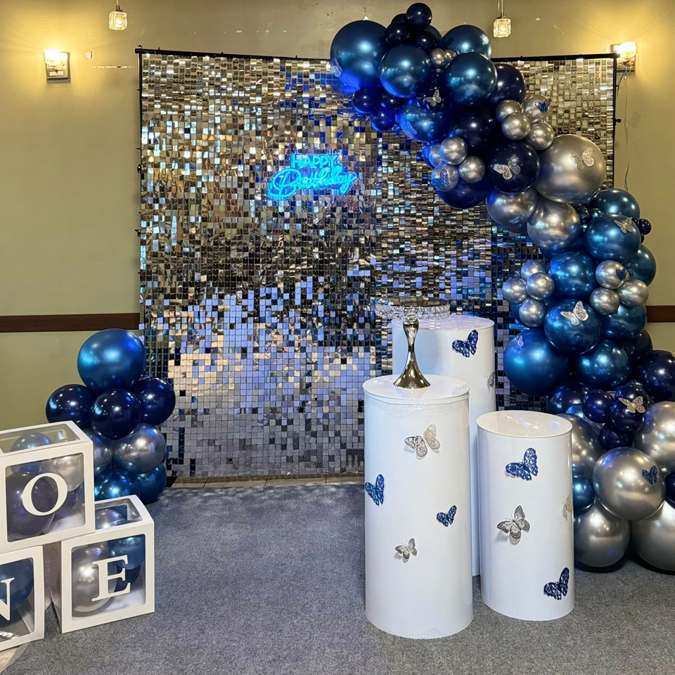A birthday party setup with blue and silver balloons, a sequin backdrop, and white pedestals.