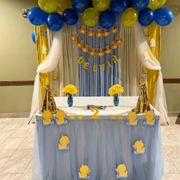 A birthday party setup with blue and yellow balloons, a "Happy Birthday Delvin" banner, and a decorated table.